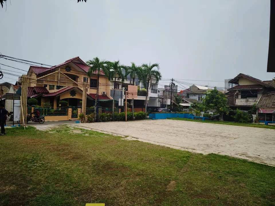 Lapangan berumput dengan deretan rumah dan pohon palem di latar belakang.