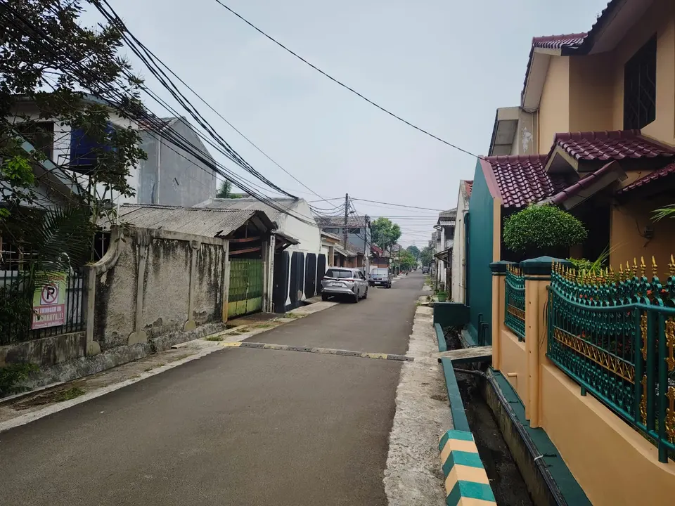 Jalan lingkungan beraspal yang diapit rumah warga dan pagar.