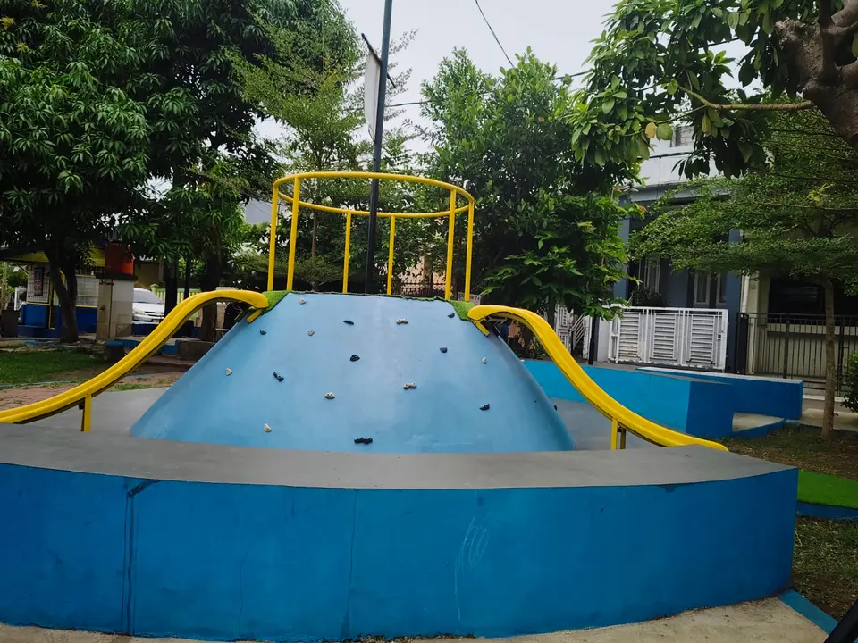 Kubah panjat biru dengan dua perosotan kuning, dikelilingi pepohonan.