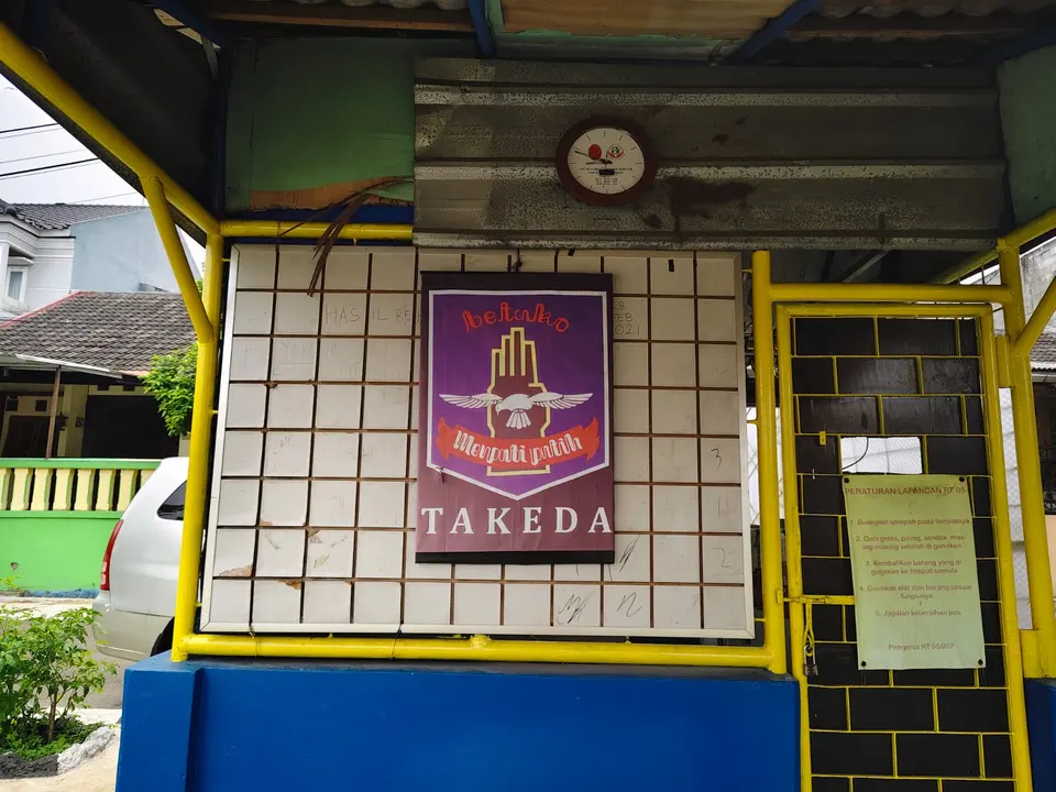 Papan bergambar Merpati Putih bertuliskan Takeda di dinding sekretariat, dibingkai pipa kuning.