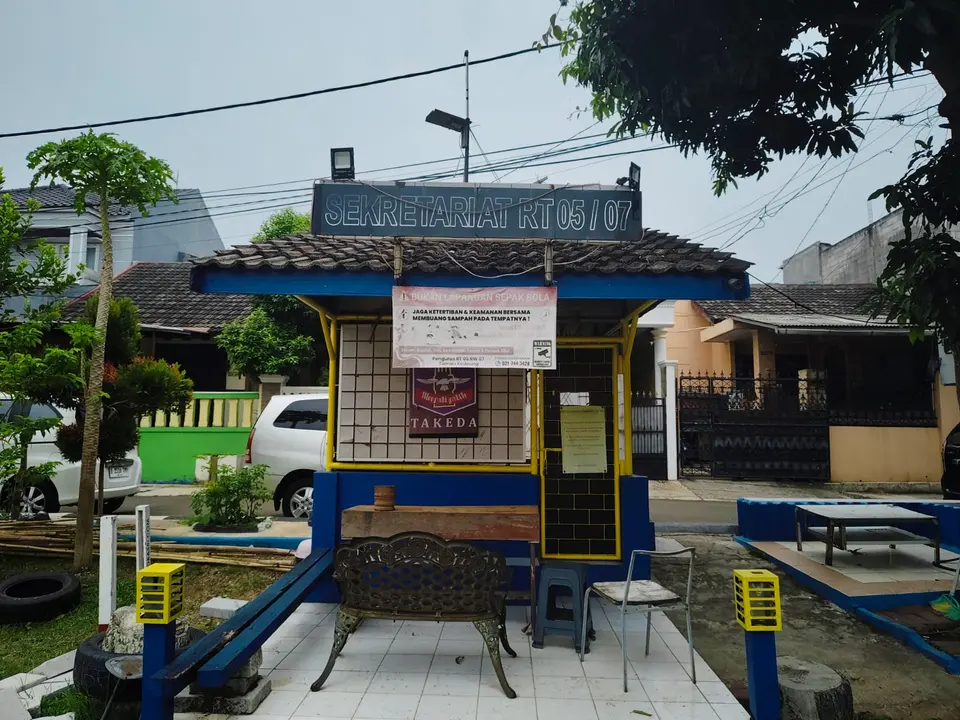Pos Sekretariat RT 05/07 berangka kuning dengan bangku dan meja di halamannya.
