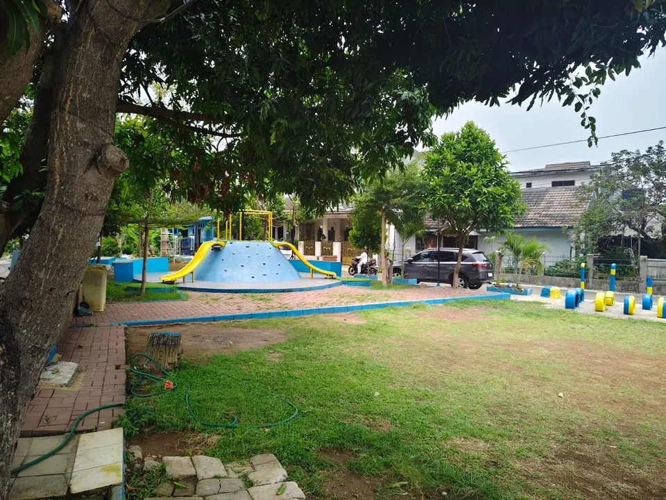 Taman bermain dengan kubah panjat biru dan perosotan kuning di bawah pohon rindang.